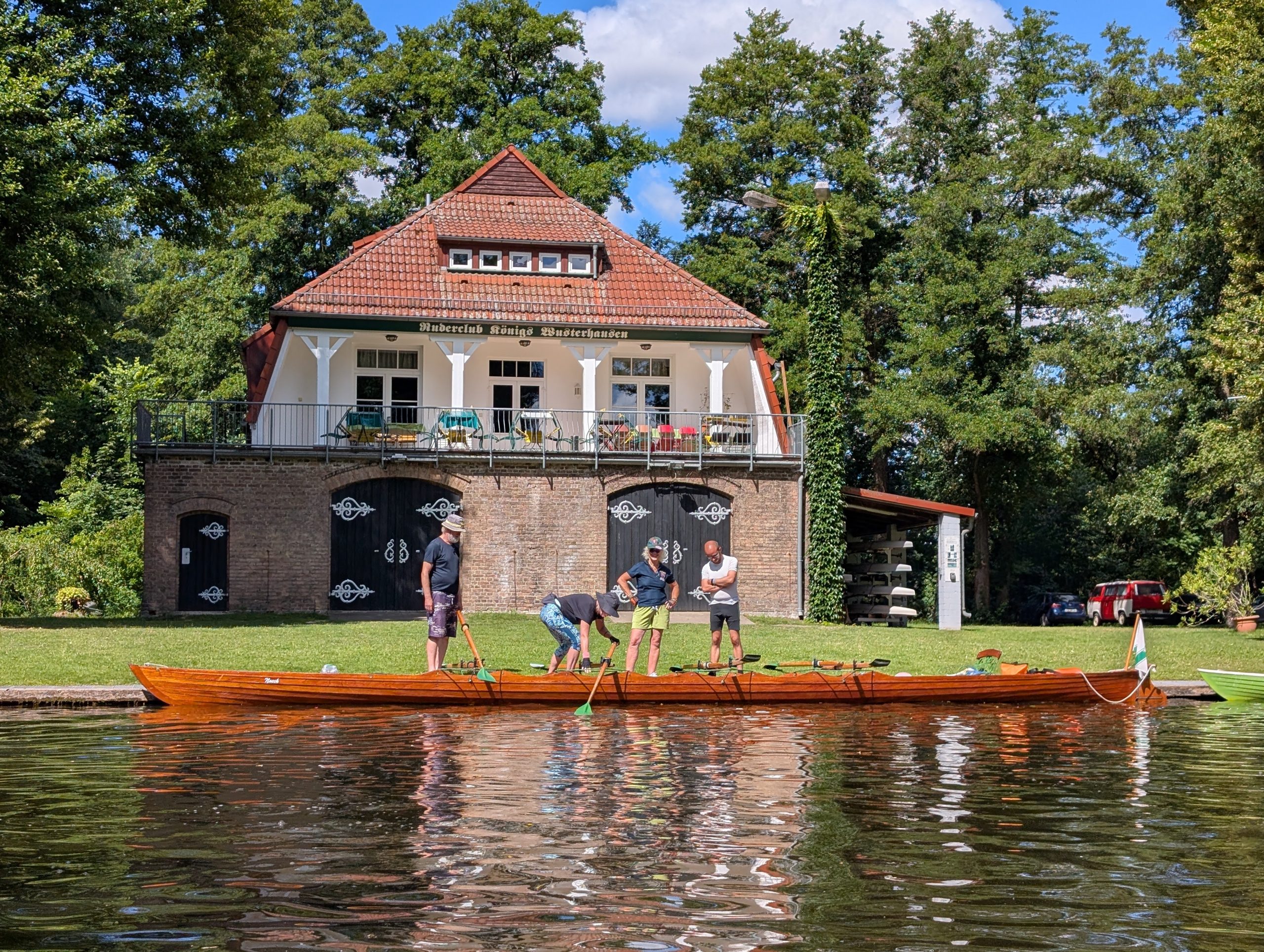 Im Vordergrund das Wasser der Dahme und ein am Steg liegender Gig-Vierer, hinter dem vier Personen stehen. Im Hintergrund das Bootshaus des Ruderclubs Königs Wusterhausen