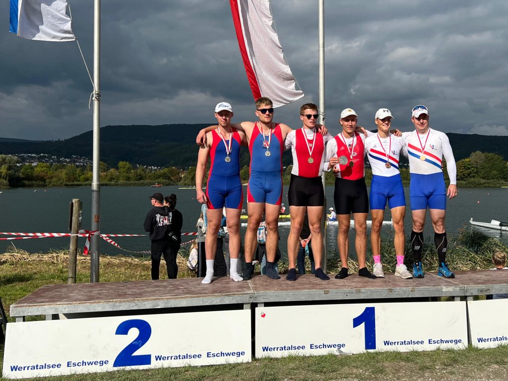 Sechs Personen stehen in Rudereinteilern und jeweils einer Medaille um den Hals auf einem Podest mit den Zahlen Zwei und Eins. Im Hintergrund sind Wasser und ein dunkel bewölkter Himmel zu sehen