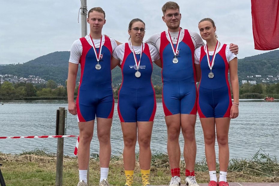Vier Personen stehen in blauroten Rudereinteilern und jeweils einer Silbermedaille um den Hals auf einem Podest mit der Zahl Zwei. Im Hintergrund ist Wasser zu sehen