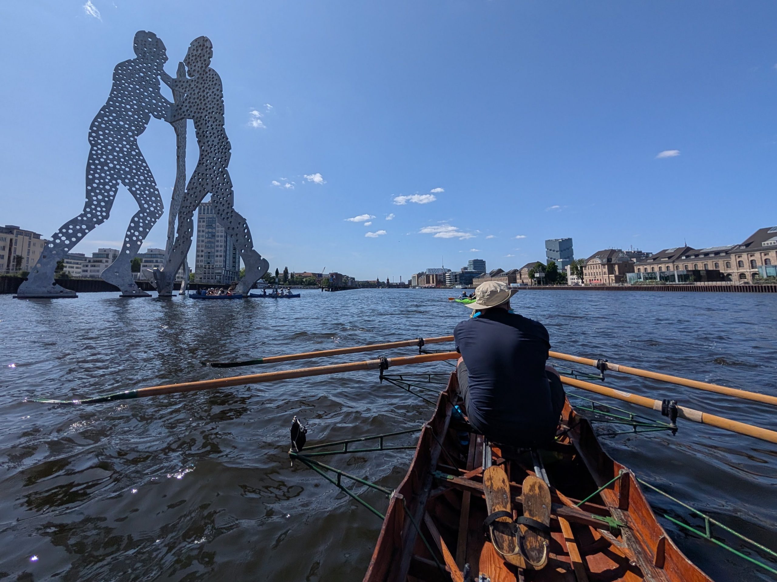 Im Vordergrund ein Gig-Vierer, der auf der Spree nach Osten fährt. Im Hintergrund die Skulptur "Molecule Man"