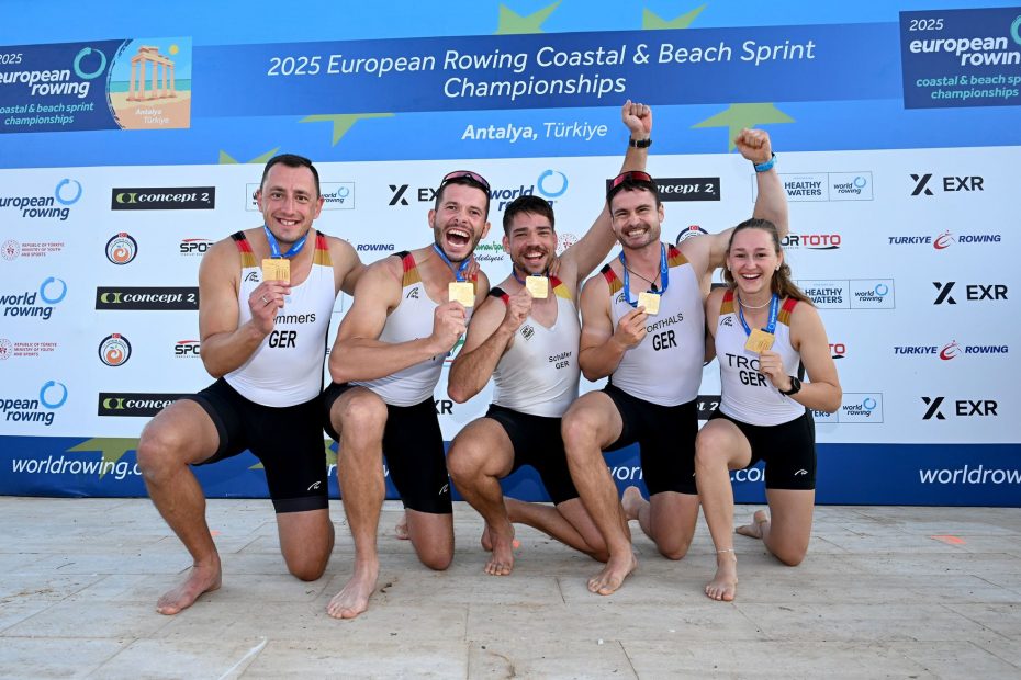 Fünf lachende Personen halten ihre Goldmedaillen in die Kamera. Im Hintergrund eine Werbewand der "2025 European Rowing Coastal & Beach Sprint Championships"