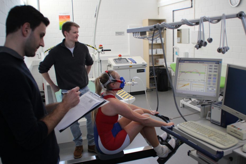 Zwei Menschen stehen neben einer Person, die auf einem Ergometer sitzt und dabei ein Atemmessgerät trägt.