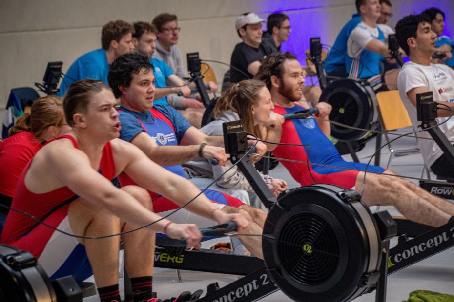Mehrere Ruderergometer, auf denen Menschen 'rudern' und dabei von anderen angefeuert werden
