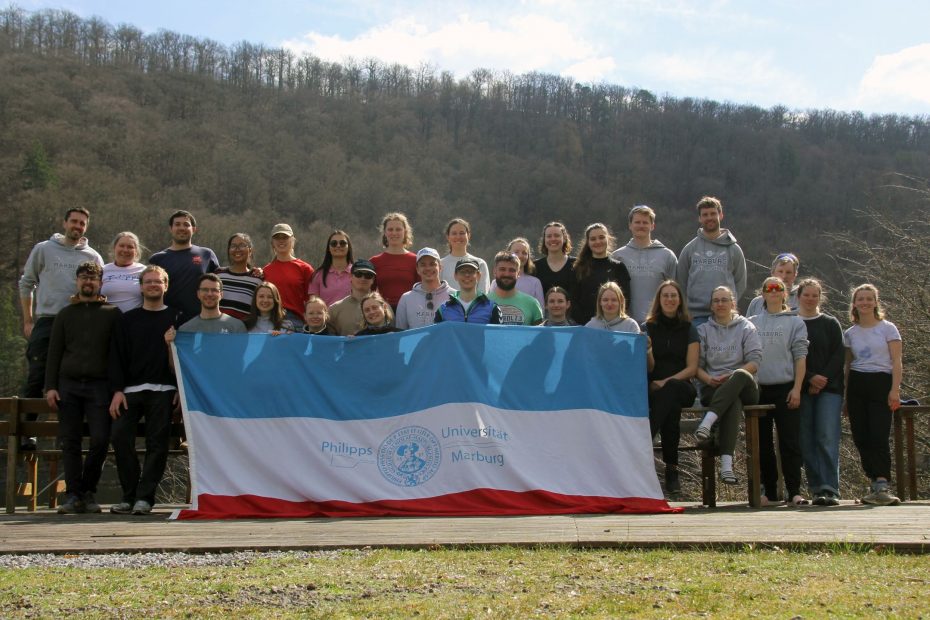 Eine Gruppe von Menschen steht hinter einer Flagge mit dem Logo der Universität Marburg, im Hintergrund ein bewaldeter Abhang