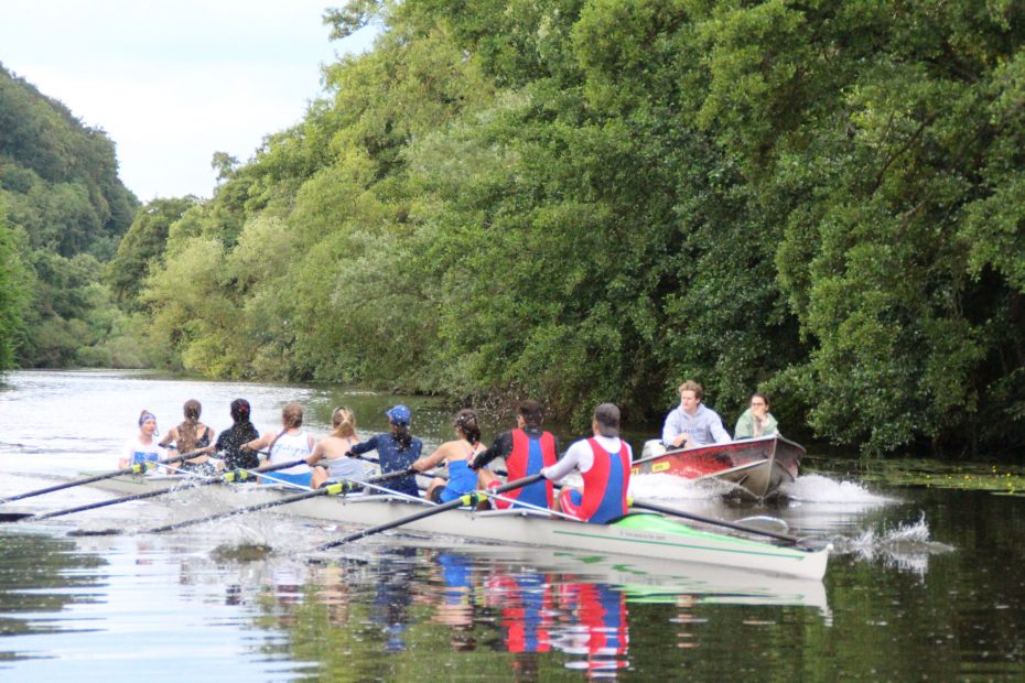 Ein Riemenachter in vollem Lauf wird von einem mit zwei Personen besetzten Motorboot begleitet.