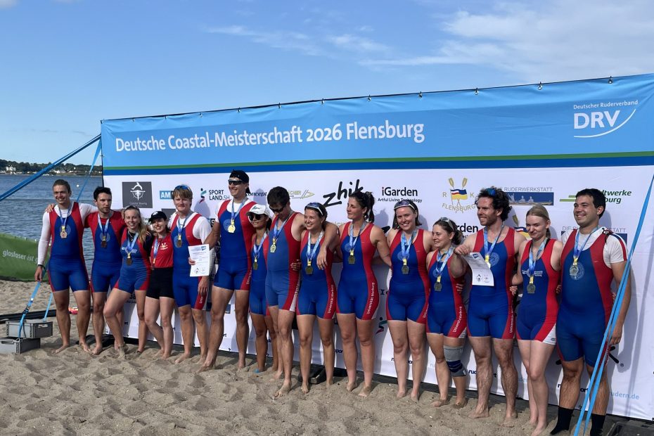 Ruder:innen posieren in blau-rot-weißen Trikots vor einem Banner mit der Aufschrift "Deutsche Coastal-Meisterschaft 2026 Flensburg"
